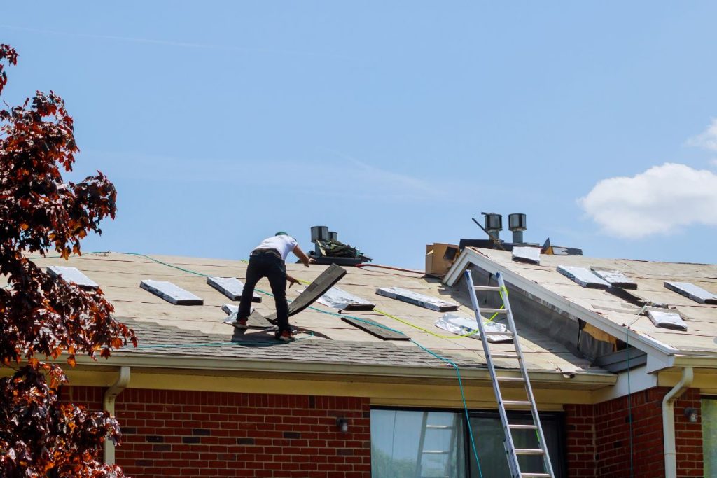 A man is repairing the roof of a house, wearing safety gear and focused on his task under a clear blue sky.