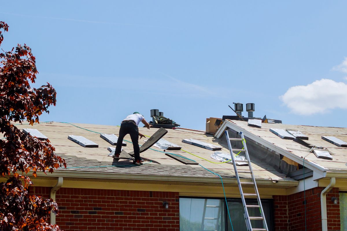 A man is repairing the roof of a house, wearing safety gear and focused on his task under a clear blue sky.