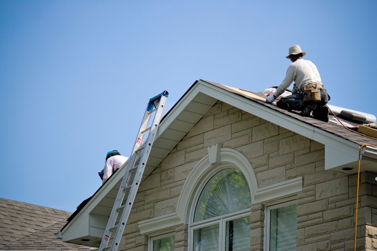 Two men installing roofing materials on a house, wearing safety gear and focused on their work.