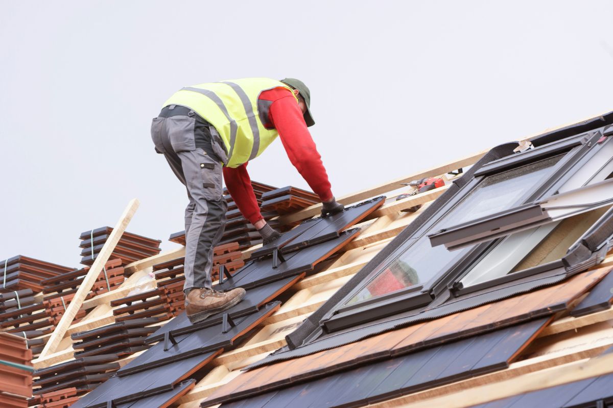 A man is repairing a roof while standing next to an open window, focused on his work.