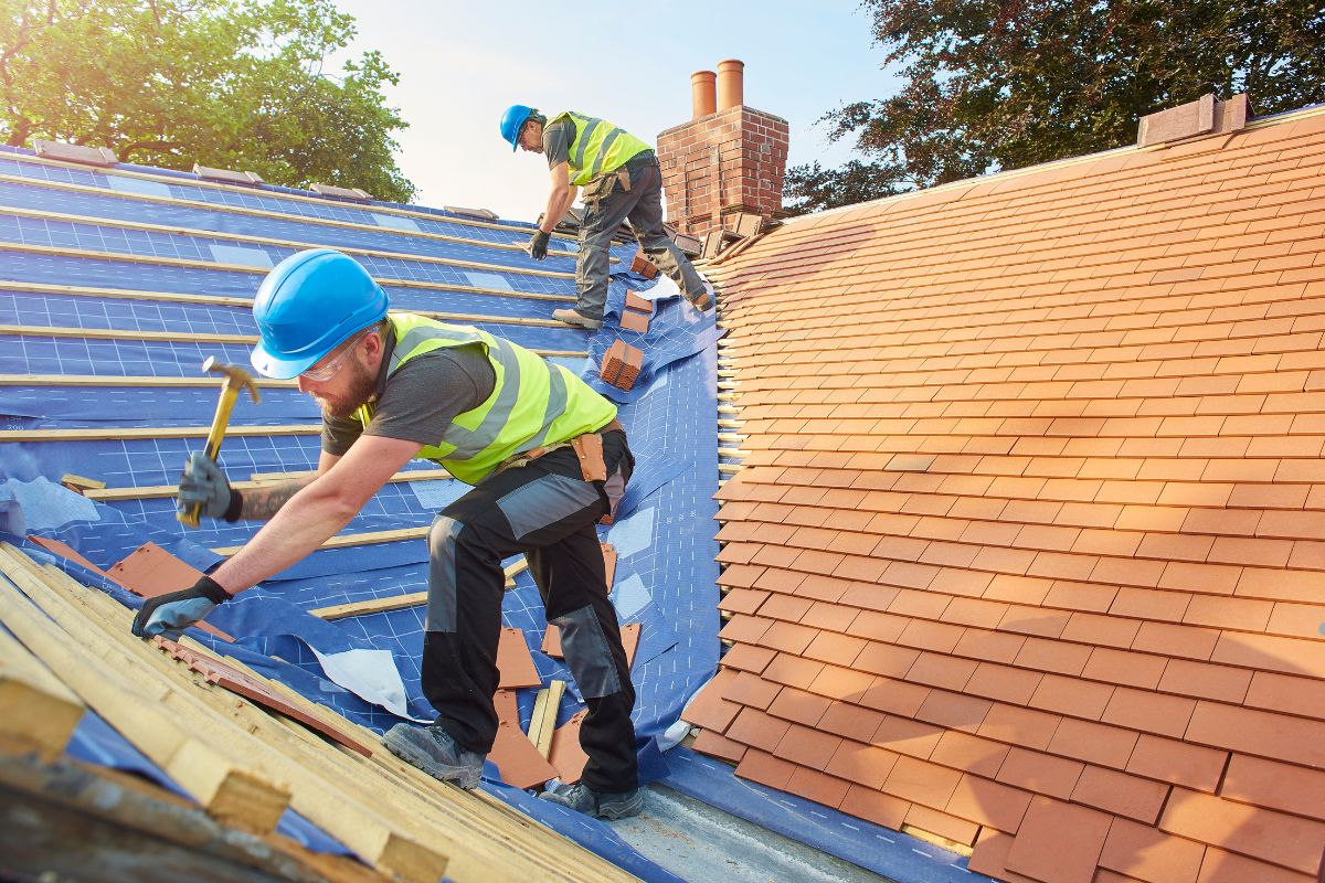 Two men using hammers to work on a roof, focused on their task under a clear blue sky.