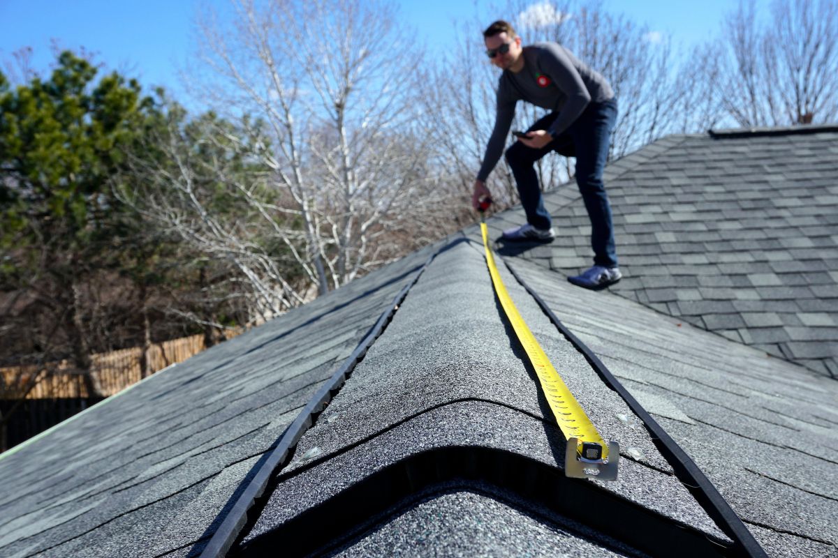 A man measuring the roof of a house with a tape measure, focused on ensuring accurate dimensions.